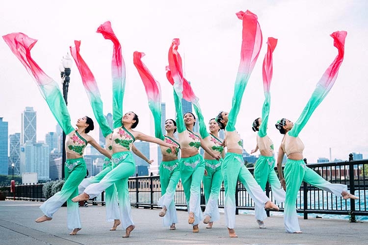 Photograph of dancers in green and pink costumes performing with long flowing ribbons in a city.