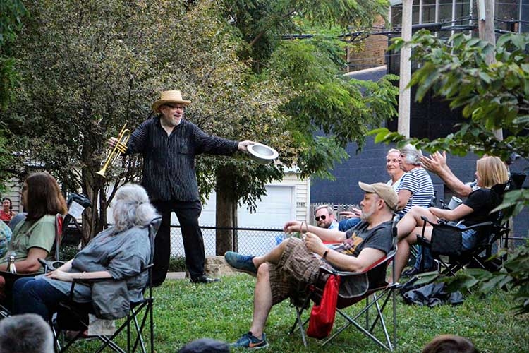 A man in a fedora holds a trumpet and bowl while performing for a seated audience outdoors.