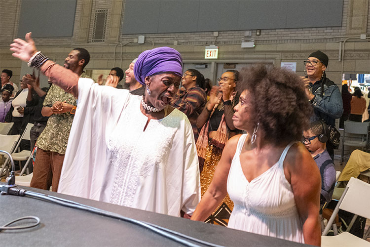 A woman in a purple headwrap and white tunic cheering in a crowd of people.
