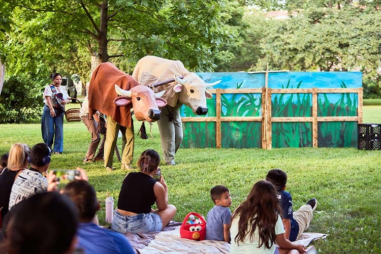 A photograph of two people in cow costumes performing outdoors for an audience on the grass.