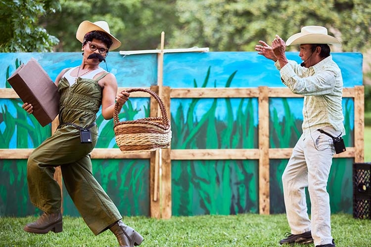 Two performers in cowboy hats act on an outdoor stage in front of a painted backdrop.