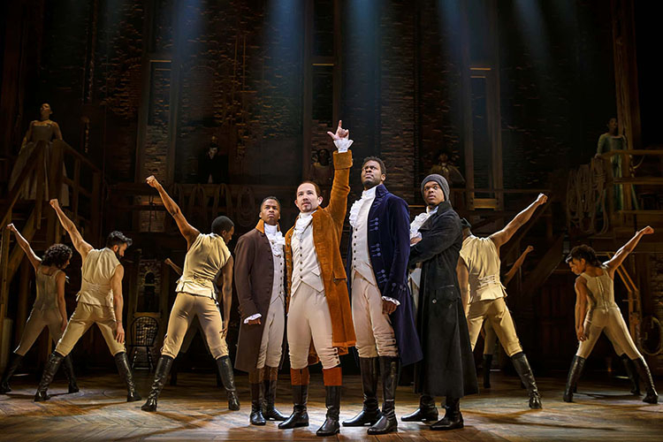 Cast members in 18th-century costumes perform on a wooden stage under spotlights in Hamilton.