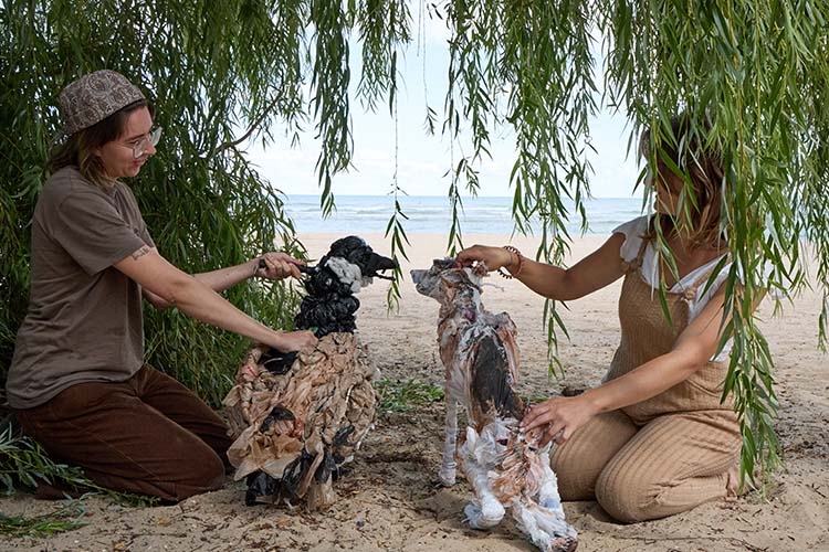 Two people kneel under a willow tree on a beach, working with puppets made of plastic waste.