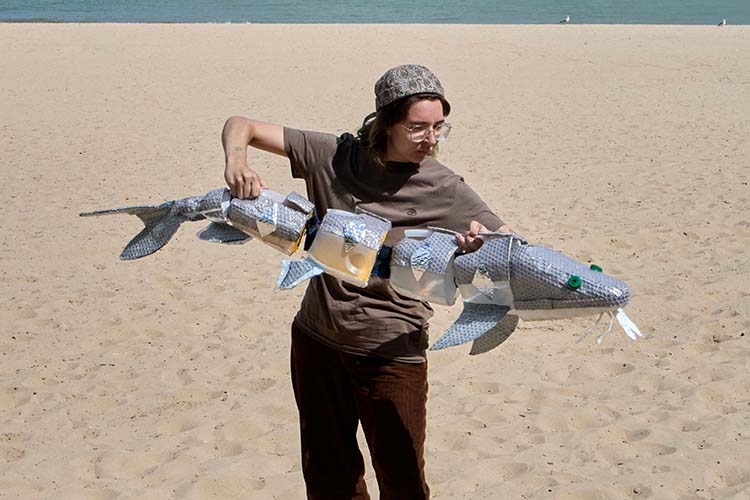 A person on a beach holds a long, segmented puppet of a fish made from recycled materials.
