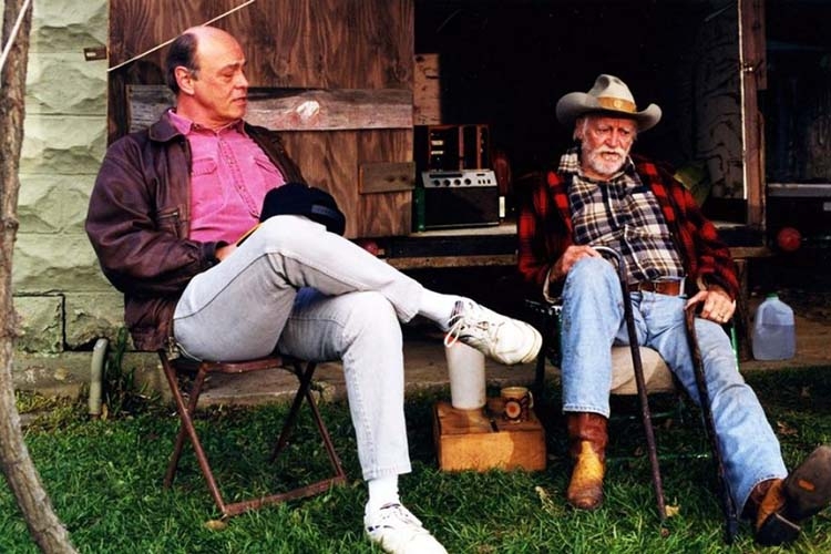 Two men sitting in folding chairs outdoors near a wooden structure.