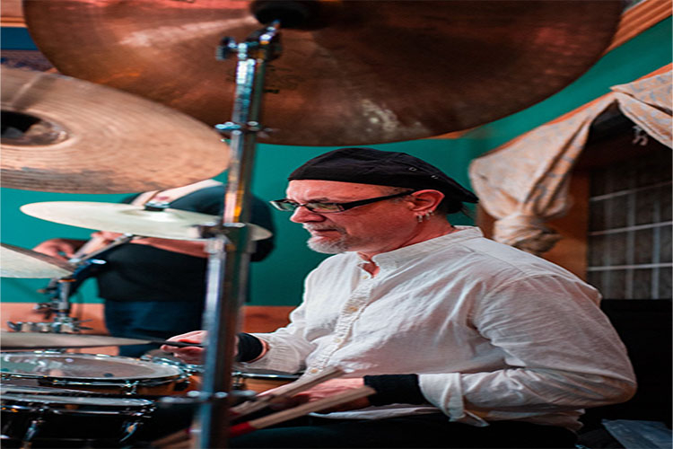 A drummer with a beard and black cap plays the drums in a room with teal walls.