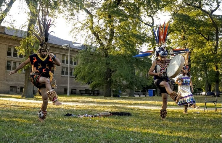 Photograph of three performers in traditional Aztec dance regalia performing on a grassy field.