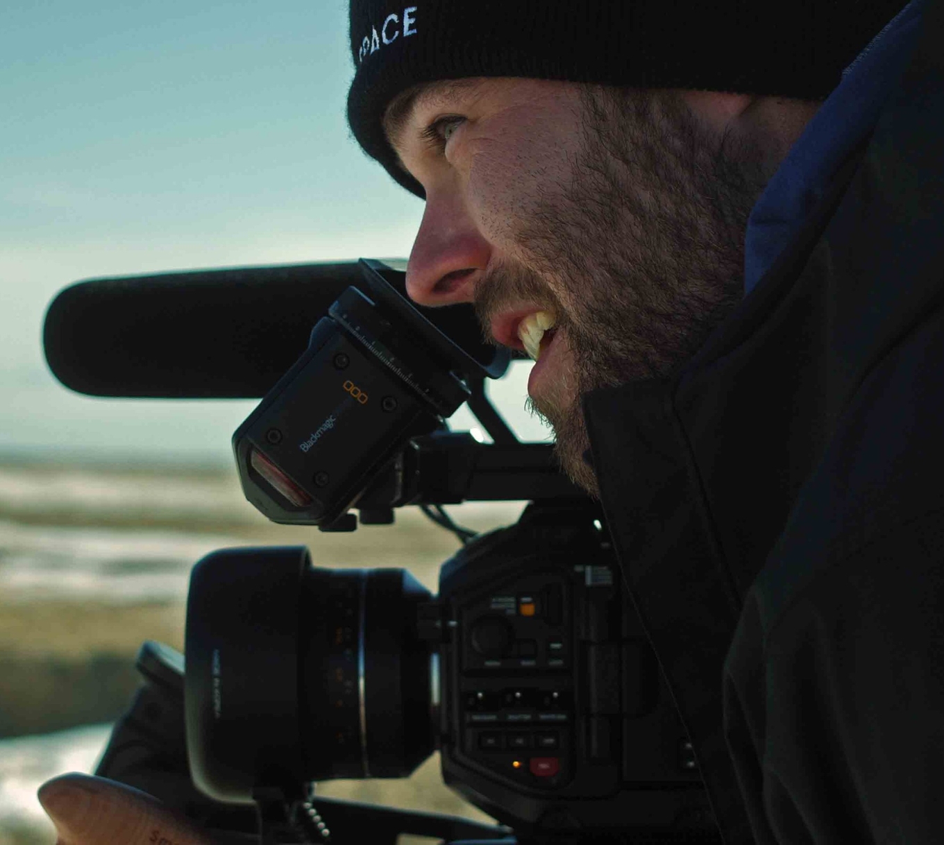 A bearded man wearing a beanie looks through a professional video camera outdoors.
