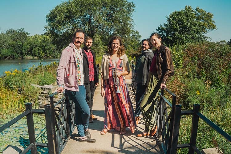 Photograph of a five-piece band standing on a wooden bridge in a sunlit park.