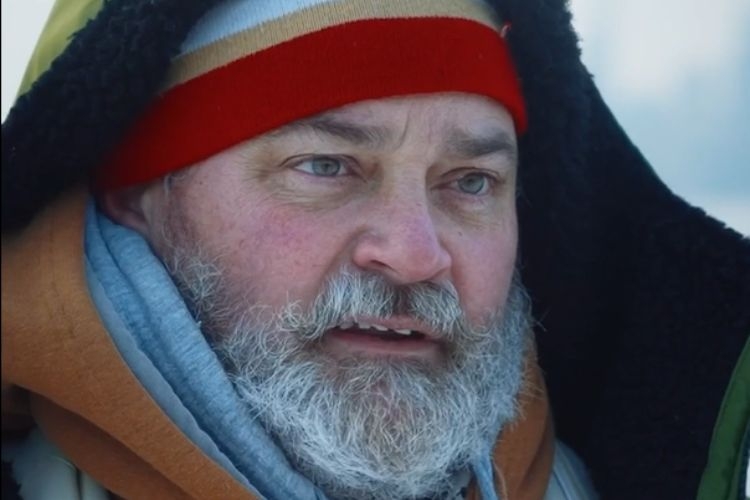 Photograph of a bearded man wearing a colorful winter hat and hooded jacket.