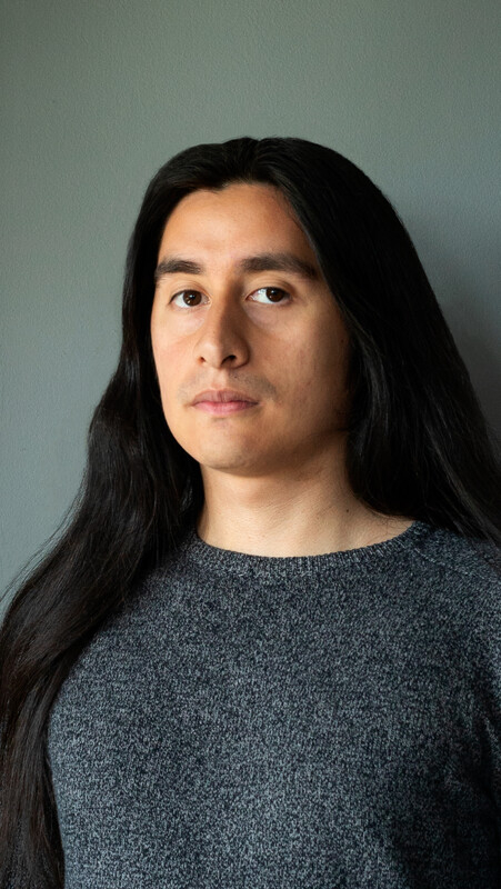 Portrait of a young man with long dark hair wearing a grey sweater against a neutral background.