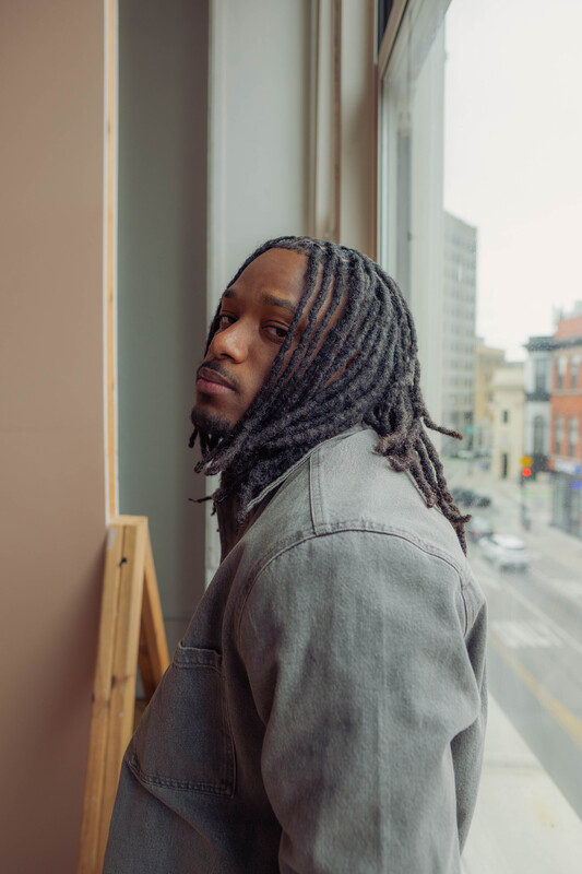A photograph of a man with dreadlocks looking back over his shoulder near a window.