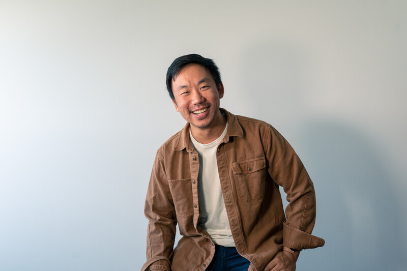 A smiling Asian man wearing a brown shirt over a white t-shirt against a light grey background.