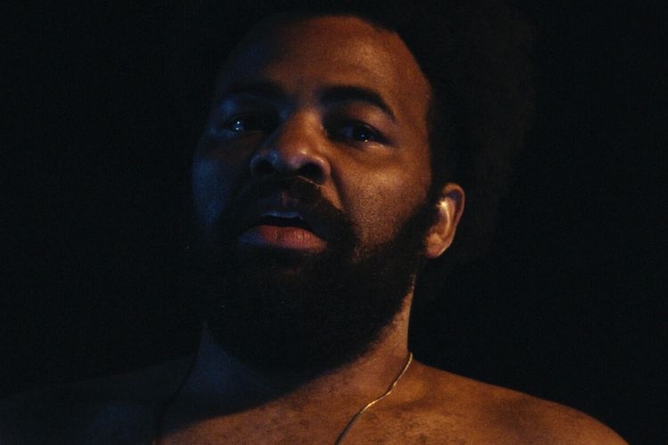 Photograph of a man with a beard and dark skin, looking up with a somber expression.