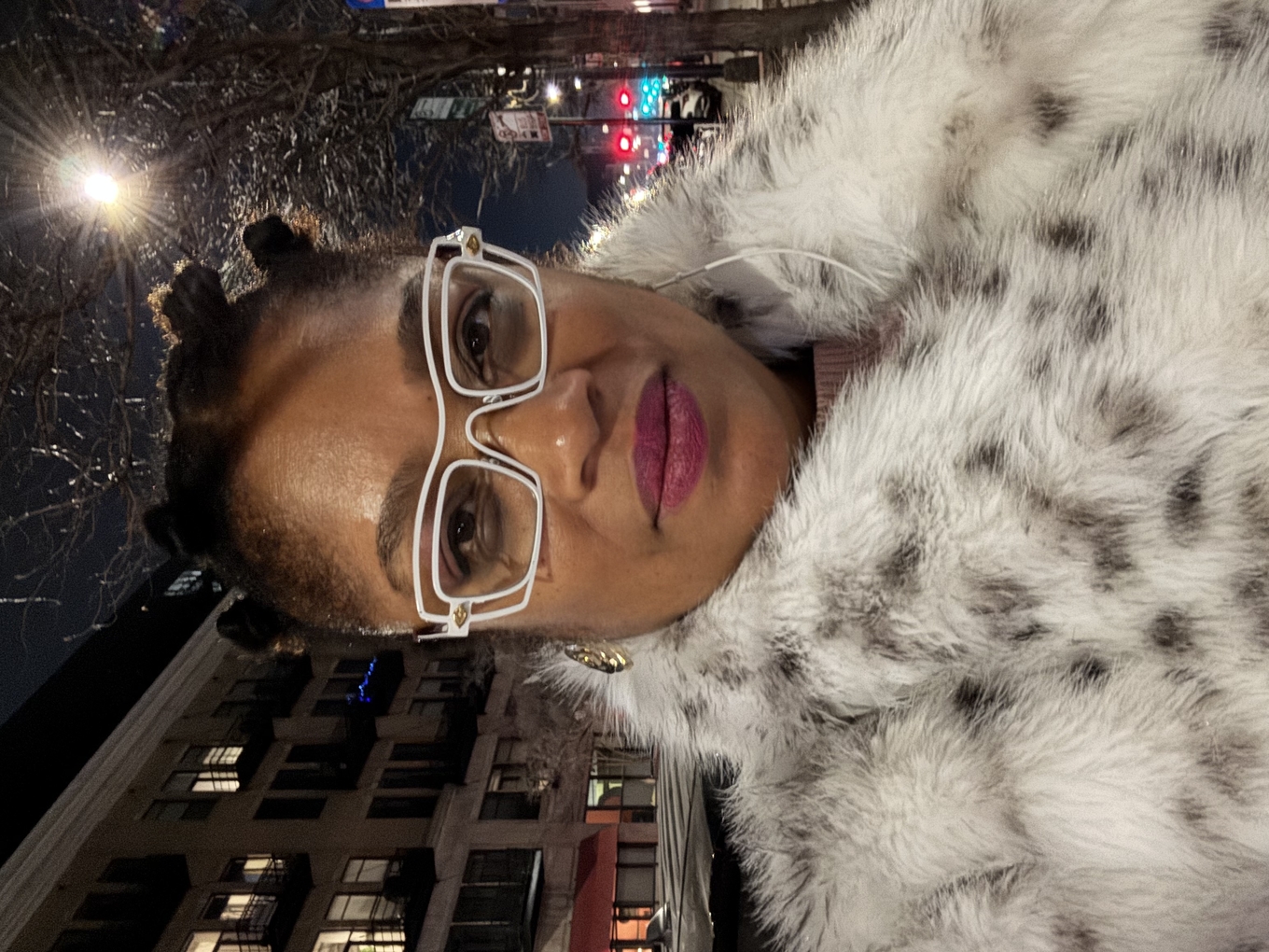 A woman with dark hair in buns wearing white-framed glasses and a faux fur coat at night.
