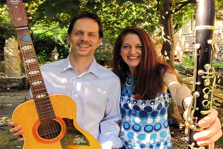 A man holding an acoustic guitar and a woman holding a clarinet smile in a garden.