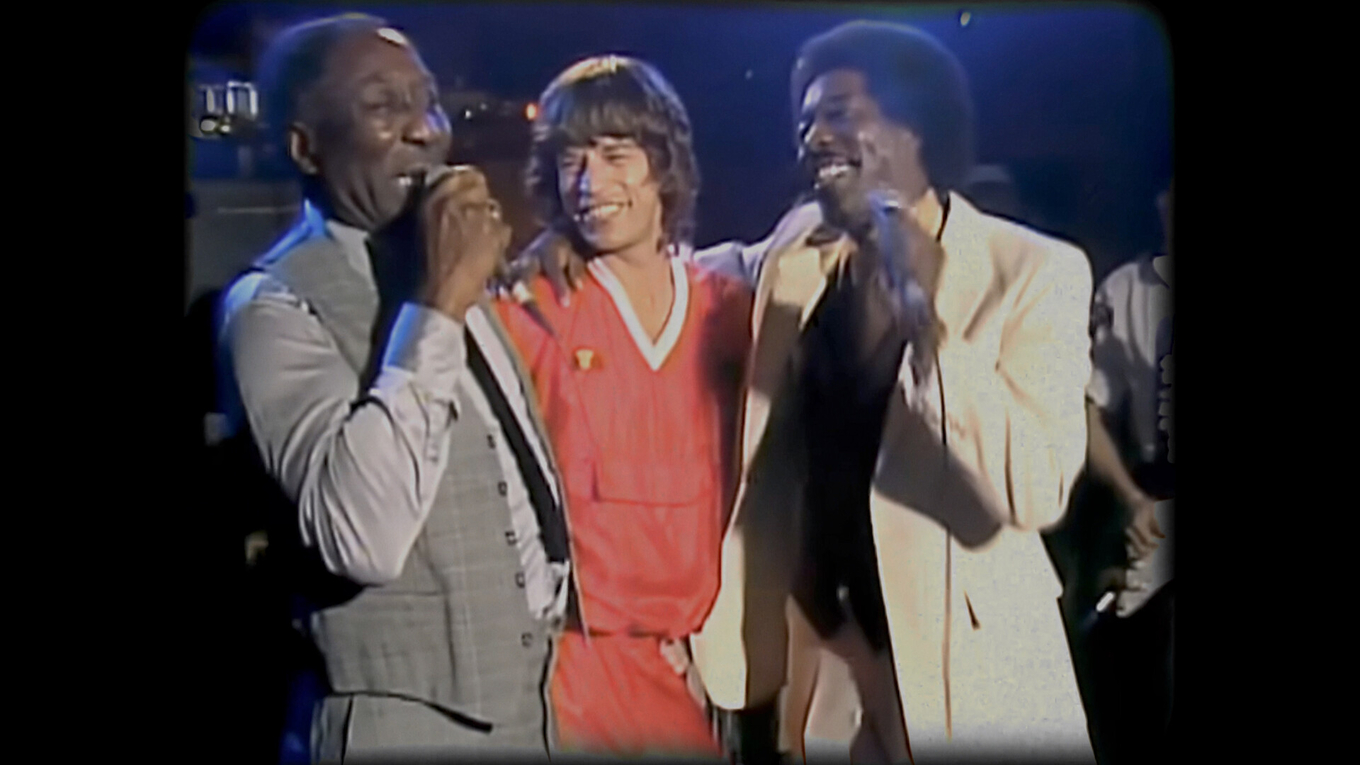 Photograph of Muddy Waters, Mick Jagger, and Buddy Guy singing together on stage.