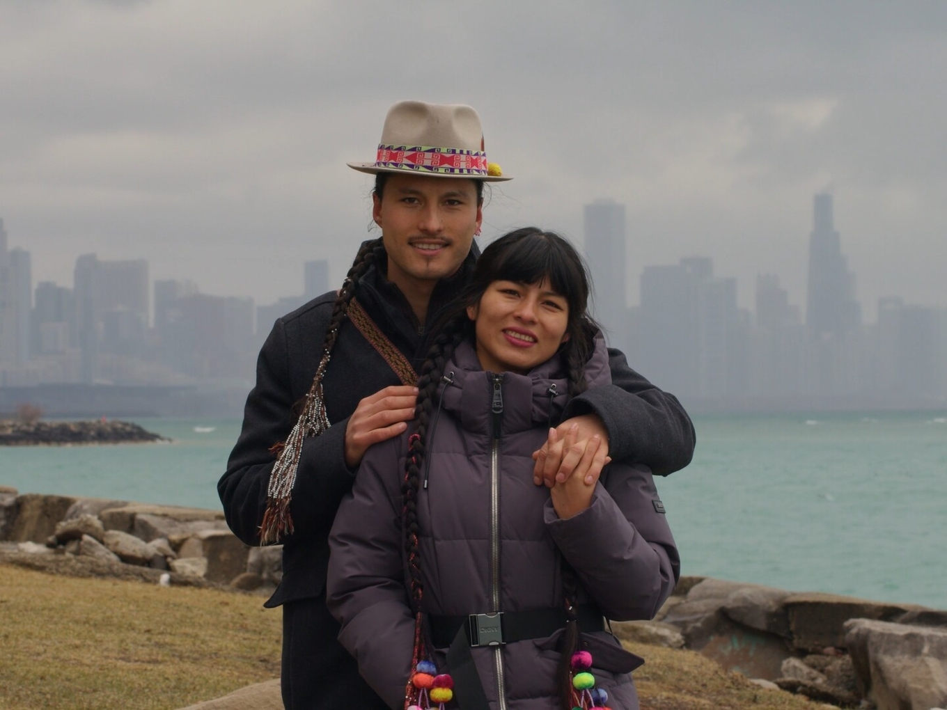 A man and woman with braids pose affectionately in front of a misty city skyline and lake.