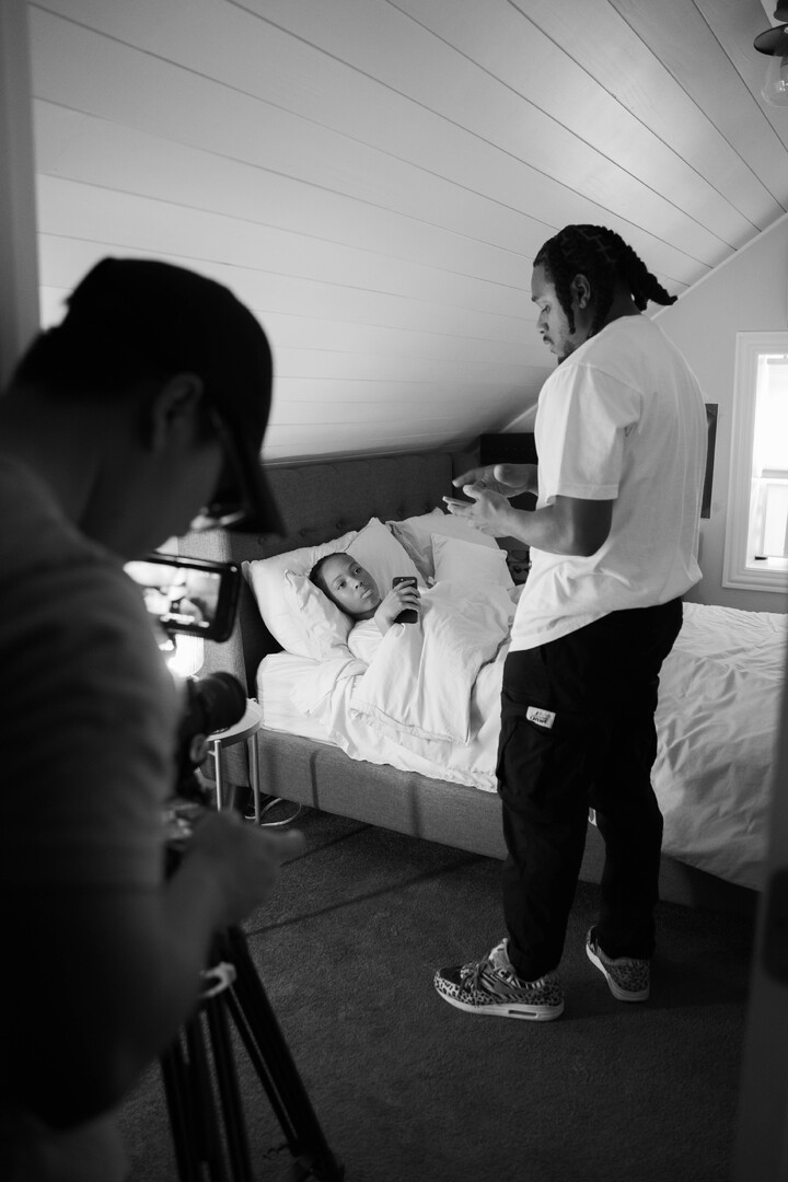 Black and white photograph of a man filming a person lying in bed holding a smartphone.