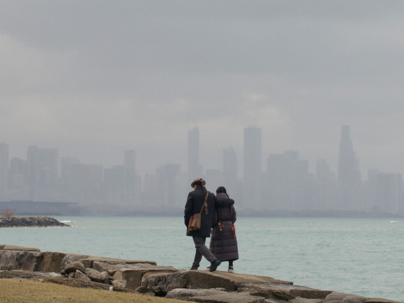 A couple walks arm in arm along a rocky shoreline with a foggy city skyline in the background.