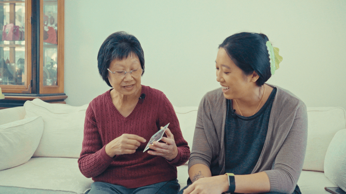 An older woman and a younger woman sitting on a white sofa and smiling while looking at a photo.