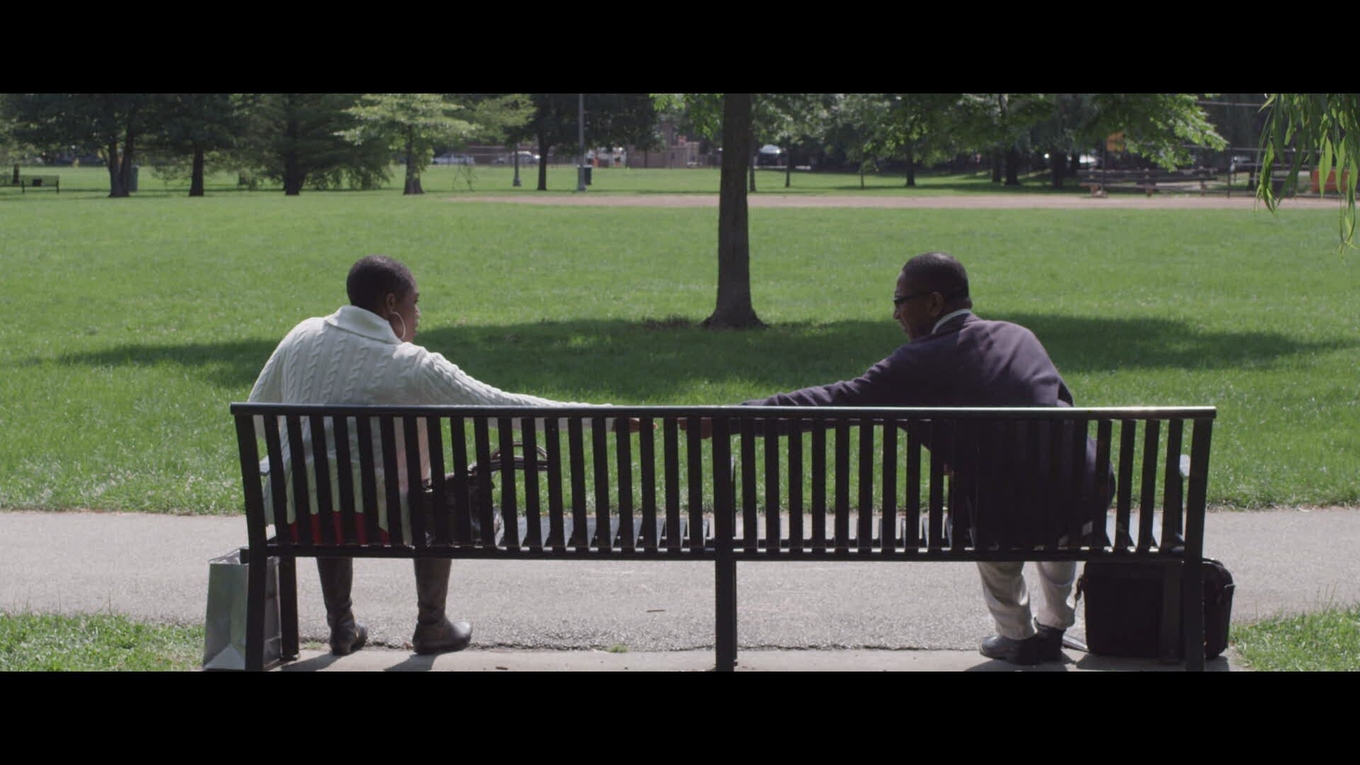 A Black couple sits on a park bench, reaching out to touch hands.