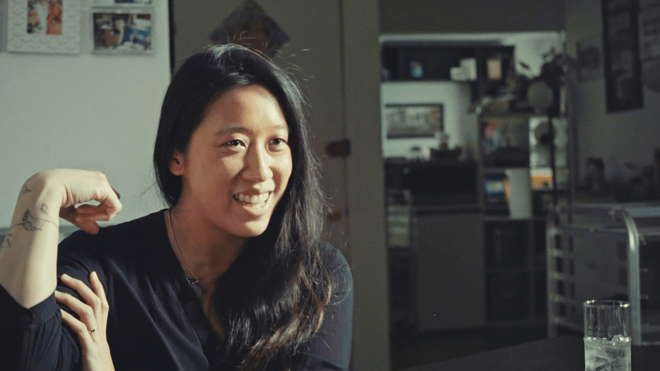 A smiling woman with long, dark hair wearing a black shirt in a dimly lit home setting.