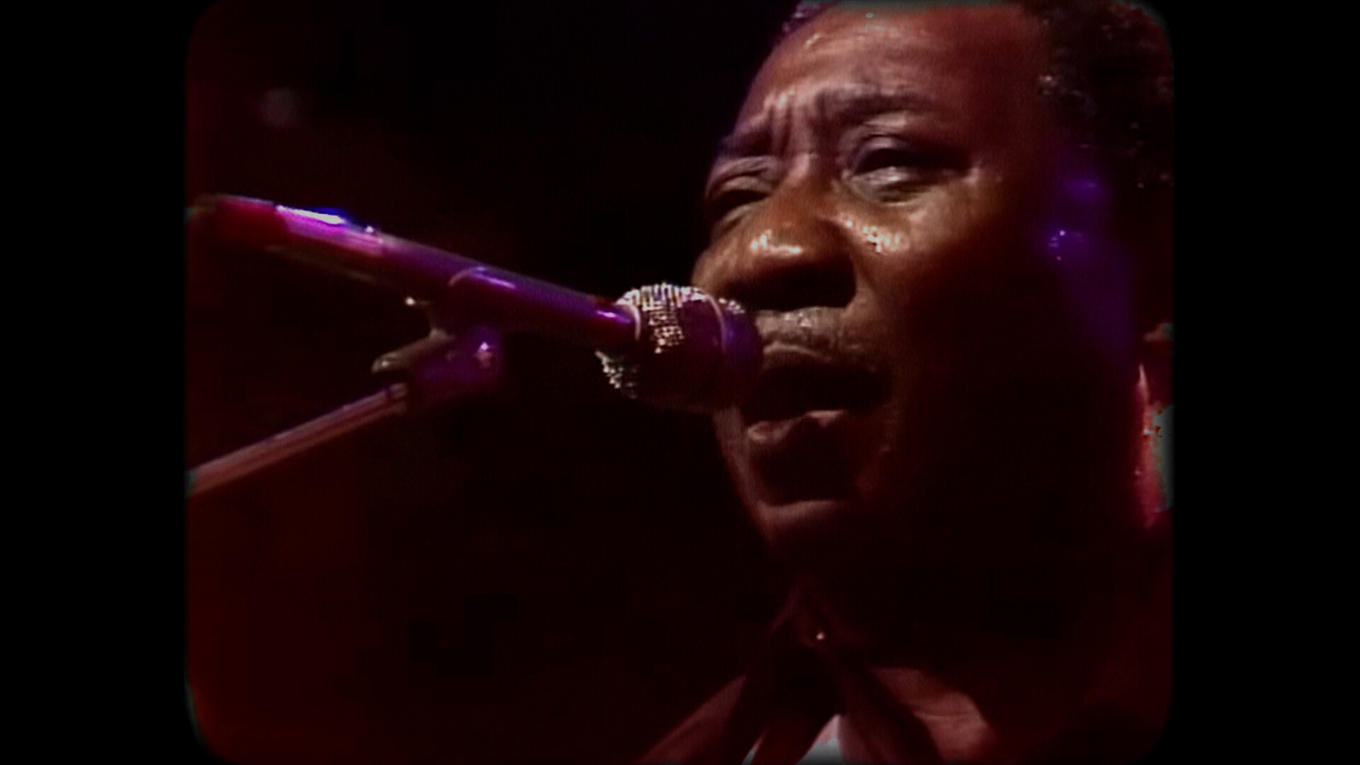 A close-up photograph of Muddy Waters singing into a microphone under low-key stage lighting.