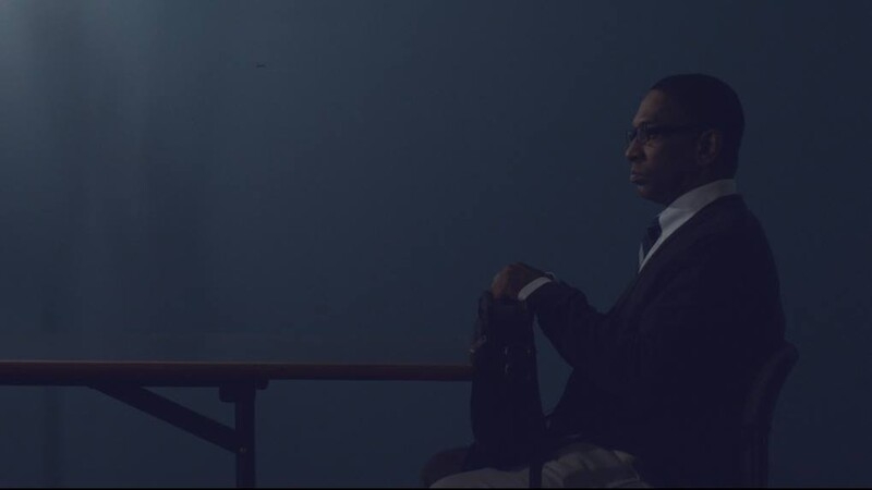 A man in a suit and glasses sits in a dimly lit, blue-toned room at a table.