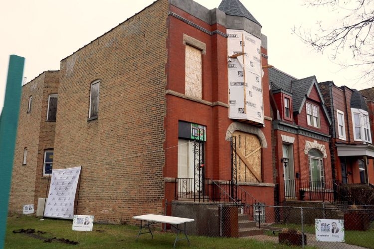 Photograph of a boarded-up red brick building under renovation with signs in the yard.