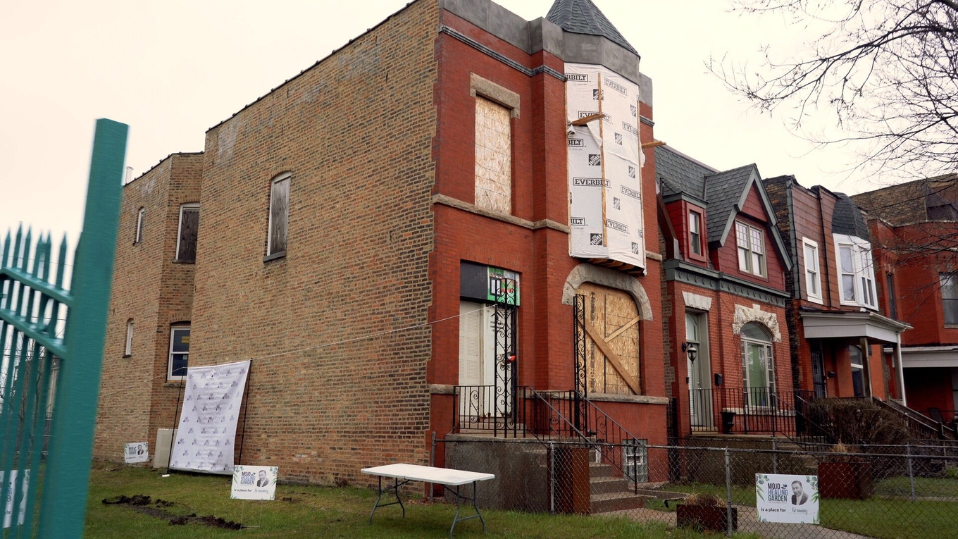 A red brick corner building under renovation with boarded windows and protective wrap.