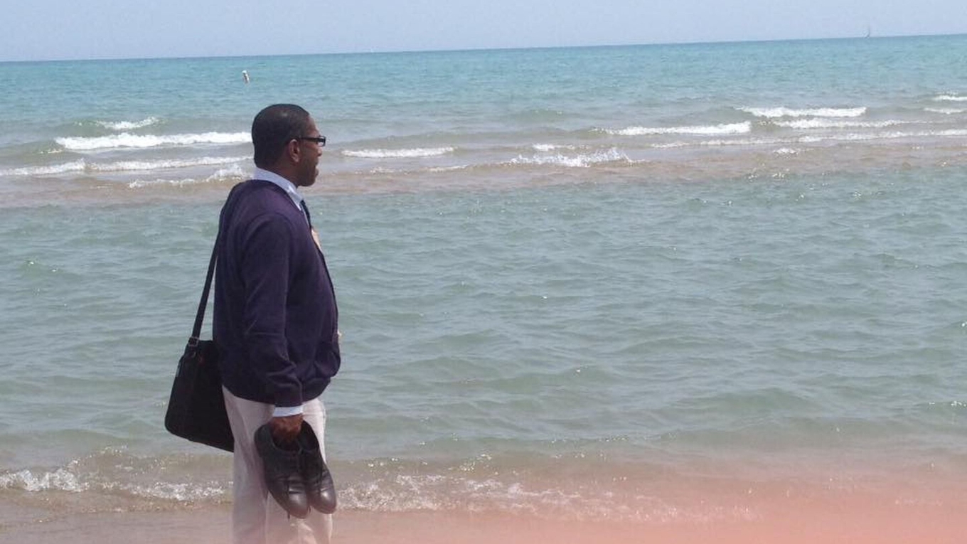 A man wearing a sweater and carrying shoes stands by the edge of the ocean, looking out to sea.
