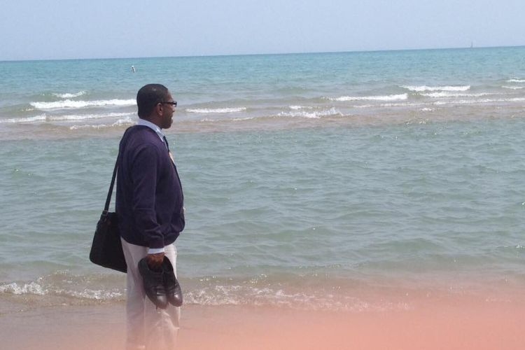 A man in a sweater and dress pants holding shoes while standing at the edge of the ocean.