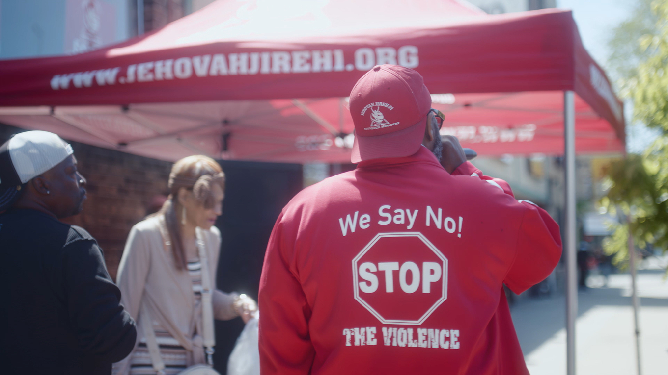 Person in a red jacket with a "Stop The Violence" sign standing before a red canopy tent.