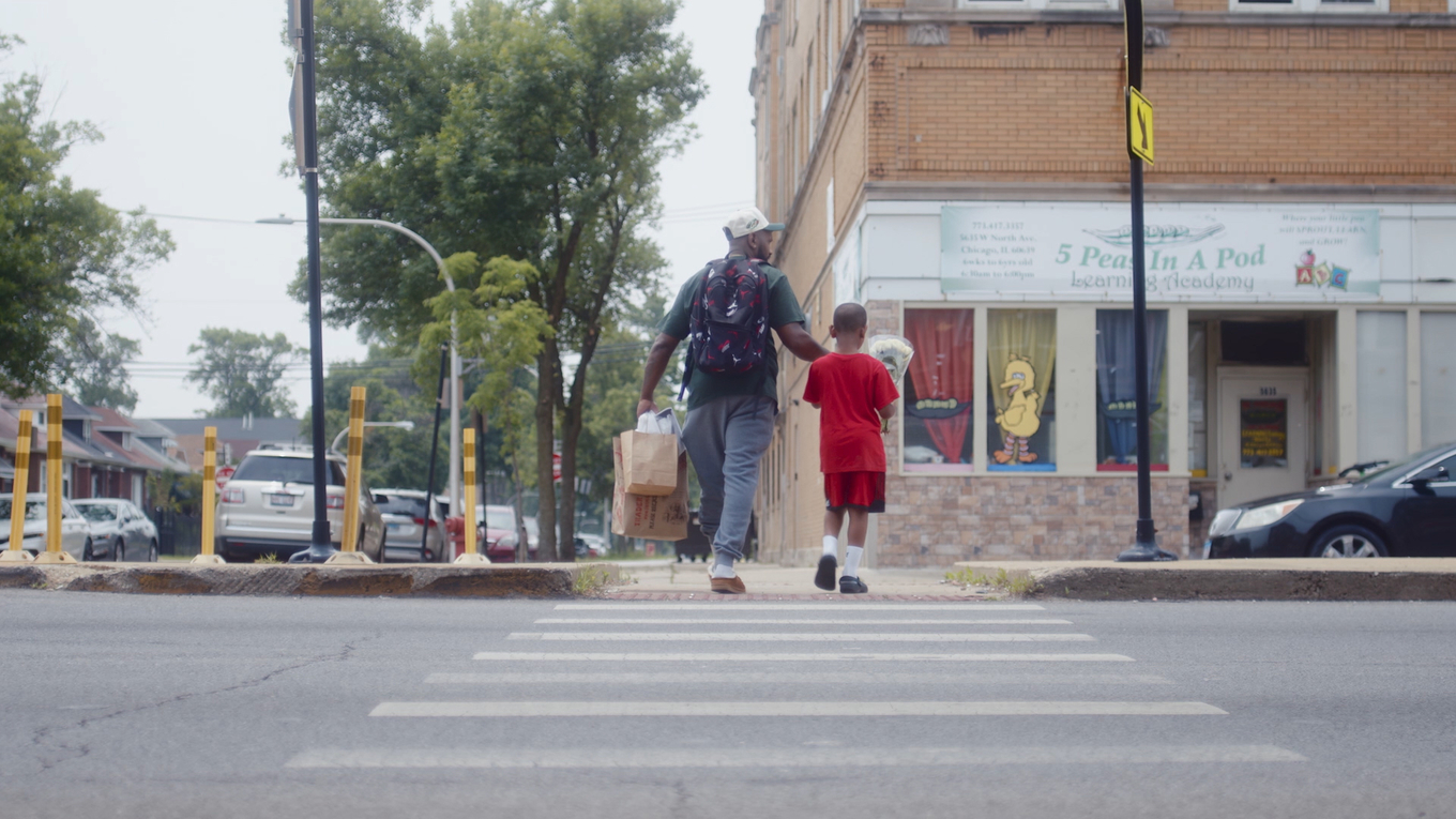 A man and a boy walk together across a city crosswalk carrying bags and flowers.