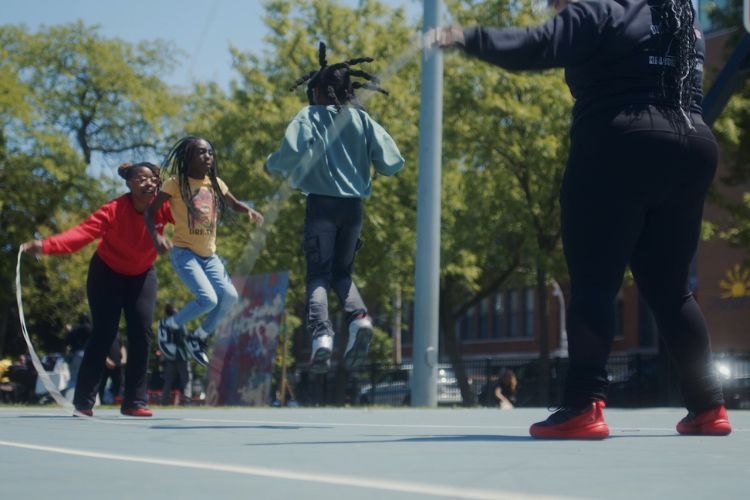 Two children jumping rope on an outdoor court, with two adults holding the ends of the rope.