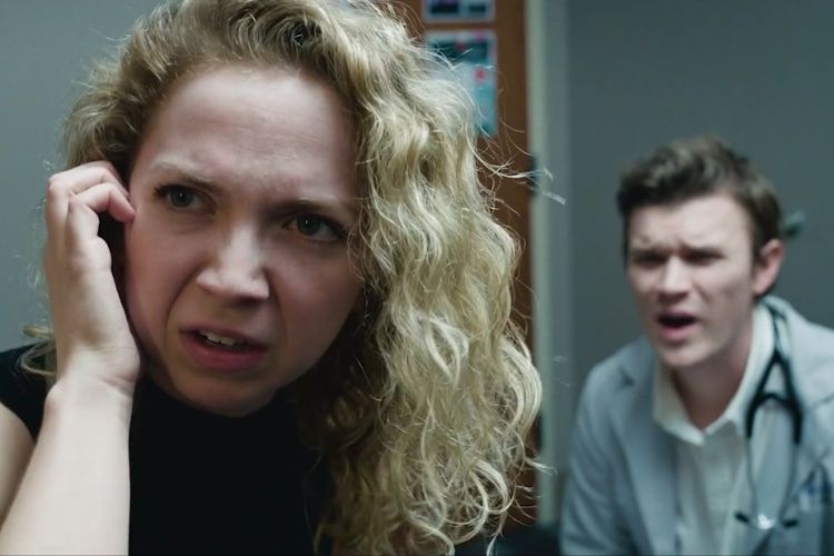 A curly-haired woman looks distressed while on a phone call, with a male doctor blurred behind her.