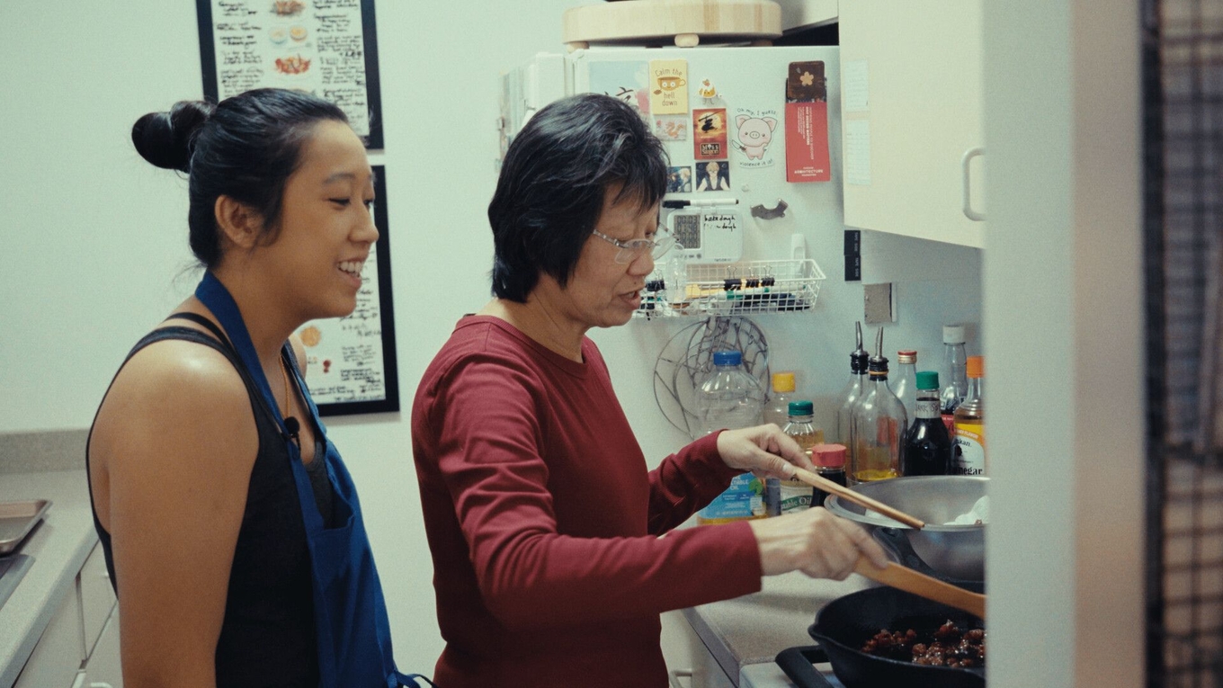A young woman watches as an older woman cooks food in a pan on the stove in a home kitchen.