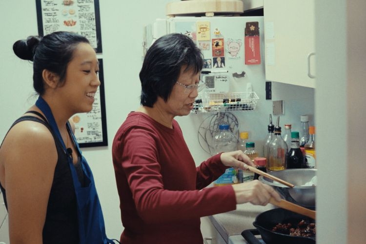 A younger woman watches an older woman cook at a stove in a home kitchen.