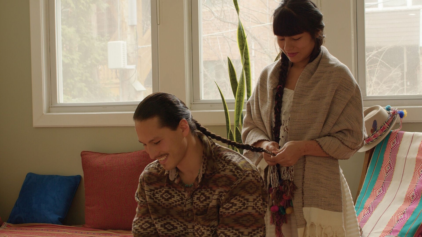 A woman braids a man's long hair in a sunlit room.