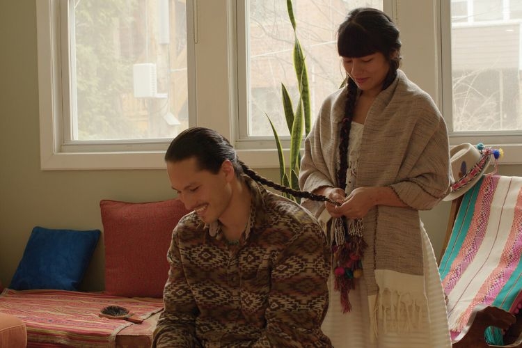 A woman braids a man's long hair in a sunlit room.
