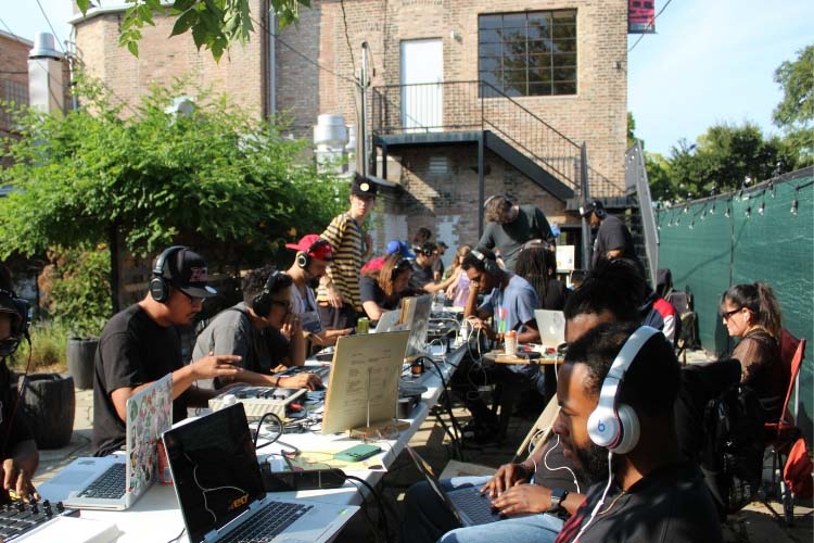 A group of people wearing headphones work on laptops at long tables in an outdoor courtyard.