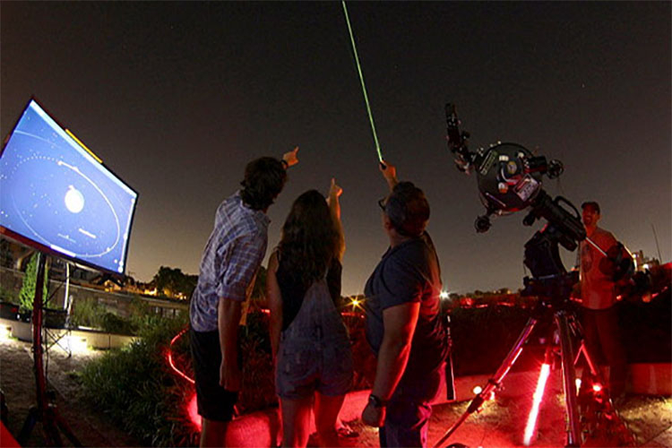 Three people standing in a field at night pointing a laser pointer at the stars near a telescope.