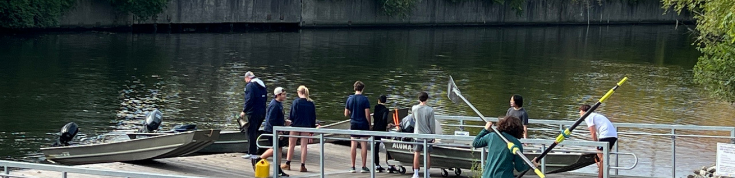 Boat Launches.png Rowing team and coaches gather at a dock with motorboats and oars.
