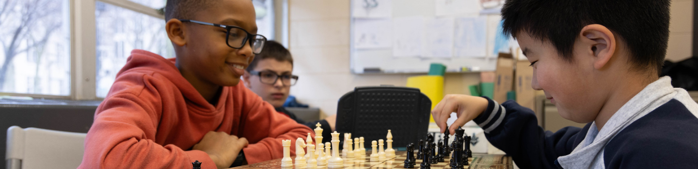 Chess Tables.png Two children play chess, another child watches.