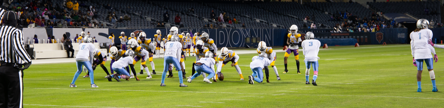Youth football game in progress. A referee watches as the offensive team, in light blue and white, prepares to hike the ball. The defensive team wears purple and gold.