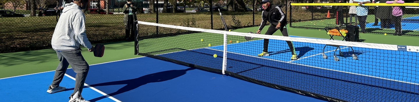 Pickleball Courts.png Two people play pickleball on a sunny day. Spectators watch from behind the fence.