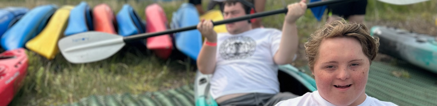 Two kayakers with Down syndrome smile near colorful kayaks and a paddle.