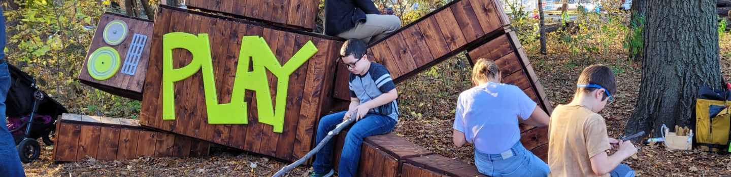 Children playing on wood play robot structure with the word “PLAY” on it in a park natue play space.