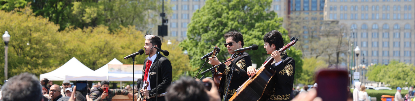 Three mariachi musicians in traditional black suits performing outdoors before a crowd.